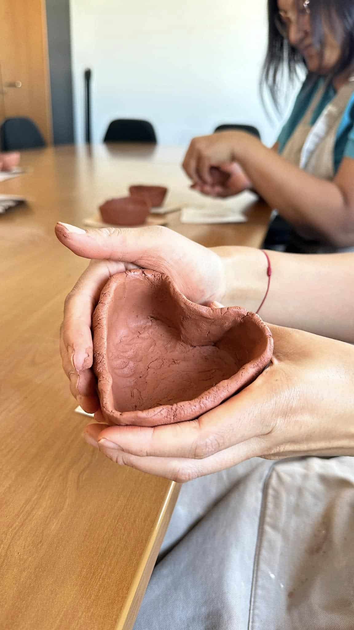Mains tenant un petit bol artisanal en terre cuite rouge en forme de cœur, lors d'un atelier de poterie.